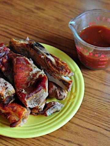 A plate of cooked western shoulder ribs with a measuring cup full of BBQ sauce