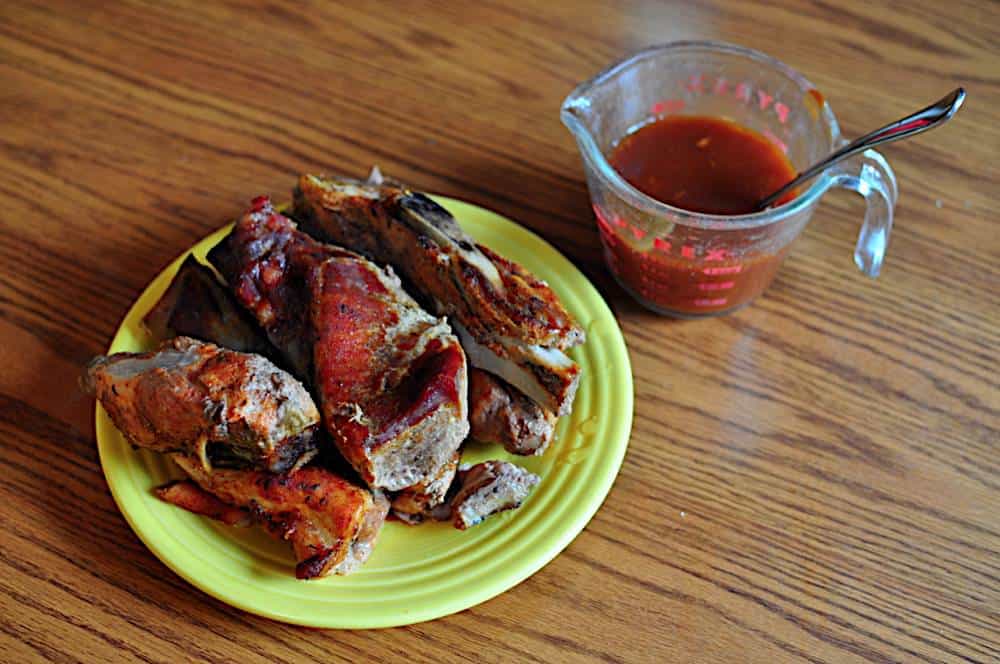 A plate of cooked western shoulder ribs with a measuring cup full of BBQ sauce