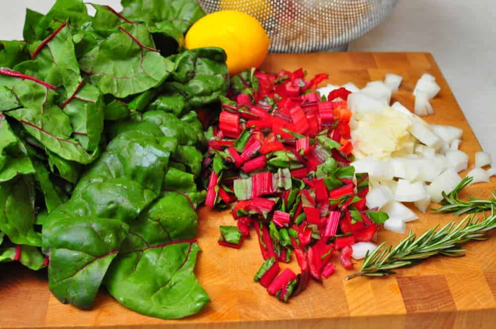 Swiss chard leaves and stems, chopped, with diced onions and a sprig of rosemary on a cutting board