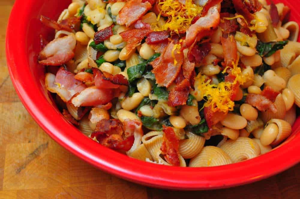 A bowl of pasta and cannellini beans, topped with chard, bacon, and lemon zest