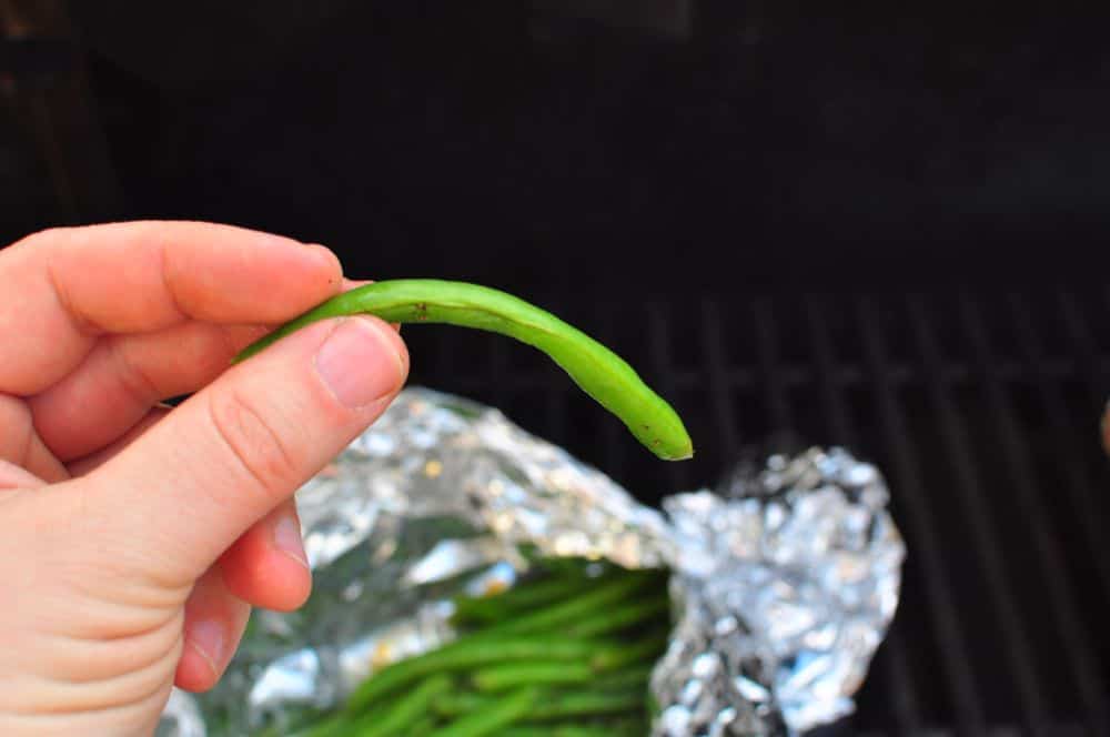 A hand holding a cooked green bean with a foil pouch of beans in the background