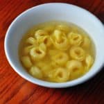 A white bowl with tortellini in broth on a reddish-brown wood background