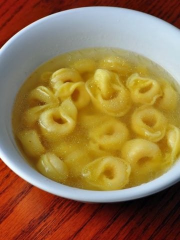 A white bowl with tortellini in broth on a reddish-brown wood background