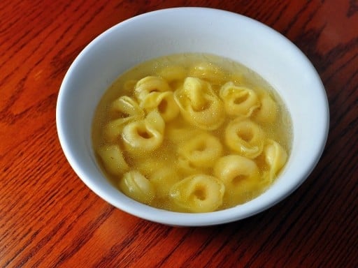 A white bowl with tortellini in broth on a reddish-brown wood background