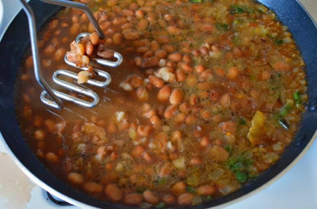 A potato masher mashing a frypan full of pinto beans and broth