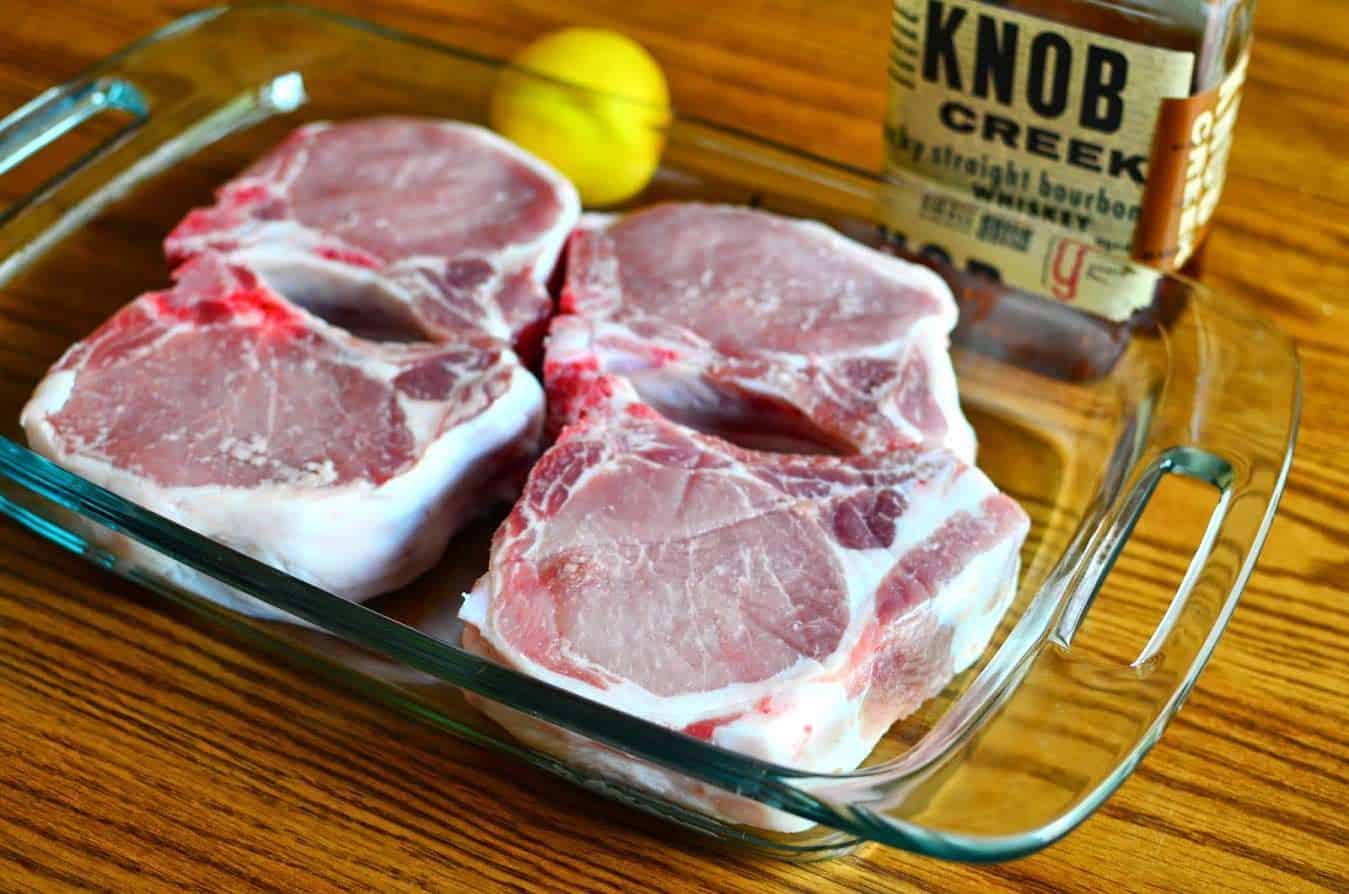 Thick pork chops marinating in a baking dish with a bottle of Knob Creek bourbon and a lemon in the background
