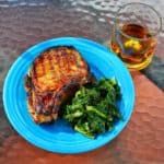 A grilled pork chop on a plate with sauteed spinach, and a glass of Knob Creek bourbon