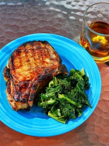 A grilled pork chop on a plate with sauteed spinach, and a glass of Knob Creek bourbon