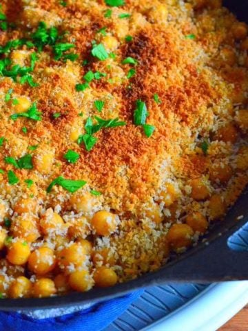 A frypan full of chickpeas, topped with a toasted bread crumb crust and a few parsley leaves