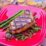 A grilled boneless pork chop with asparagus and a bed of kale on a red plate