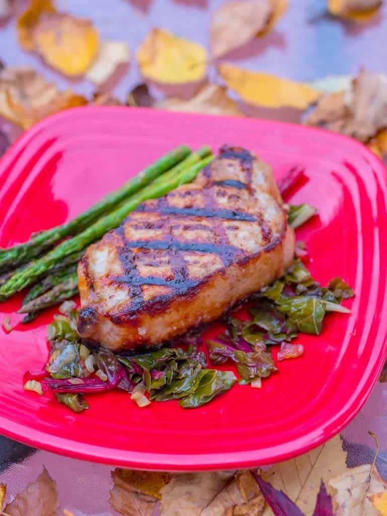 A grilled boneless pork chop with asparagus and a bed of kale on a red plate