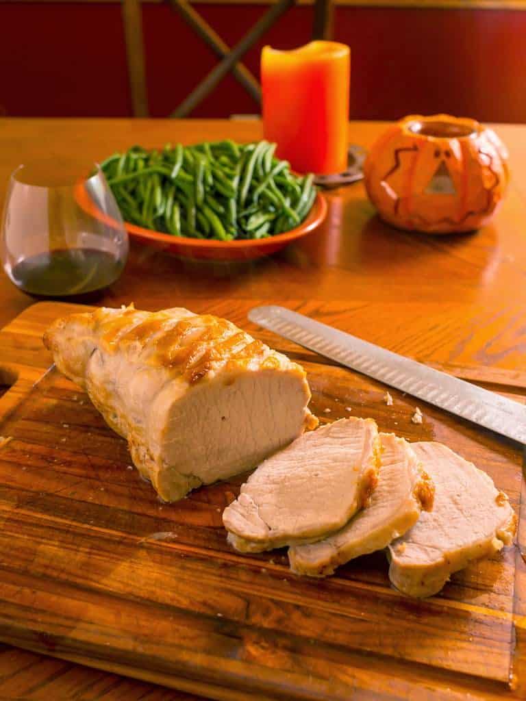 A sliced pork loin roast on a cutting board with a carving knife, green beans, a candle, and a pumpkin in the background