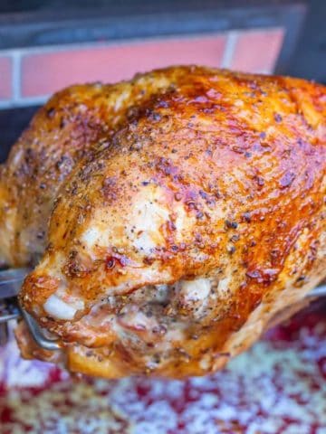 A turkey breast on a grill's rotisserie, with a lit rotisserie burner in the background.