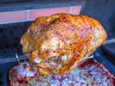 A turkey breast on a grill's rotisserie, with a lit rotisserie burner in the background.