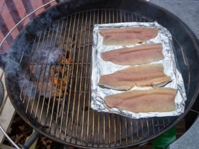 Kettle grill with coals and smoking wood on one side and a foil tray of uncooked trout filets on the other side