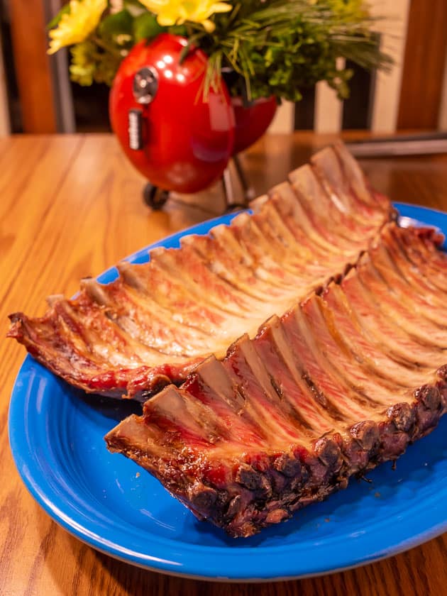 Two cooked slabs of ribs on a platter in front of a flower arrangement that looks like a kettle grill