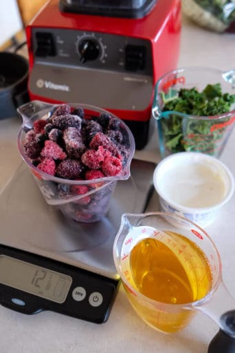 Apple juice, frozen berries on a scale, yogurt, kale and a red Vitamix blender