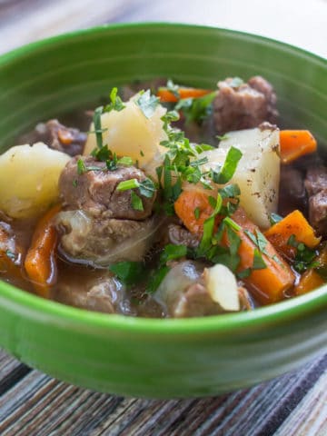 Side view of a green bowl full of stew, with chunks of potatoes, carrots, and lamb, sprinkled with parsley.