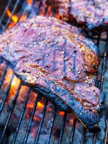 Ribeye steak on a grill, over charcoal, with a flare of fire and a second ribeye in the background