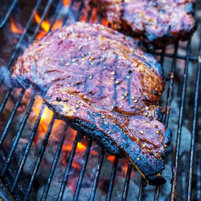 Ribeye steak on a grill, over charcoal, with a flare of fire and a second ribeye in the background