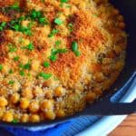 A frypan full of chickpeas, topped with a toasted bread crumb crust and a few parsley leaves