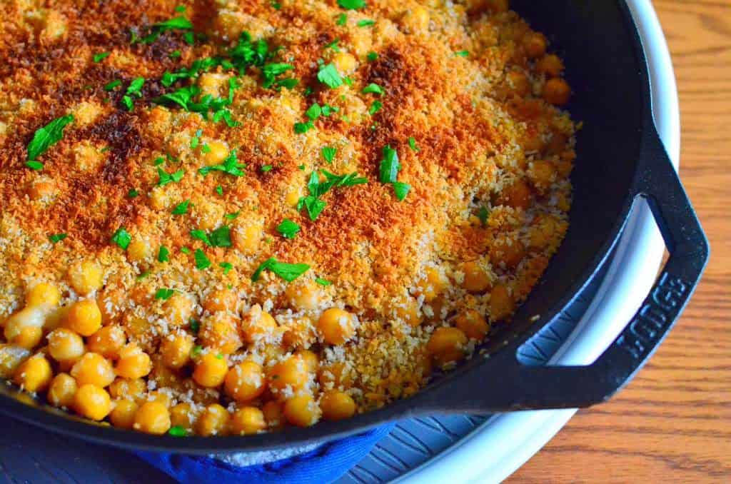 A frypan full of chickpeas, topped with a toasted bread crumb crust and a few parsley leaves