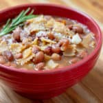 A red bowl of Venetian pasta and beans garnished with a sprig of rosemary.