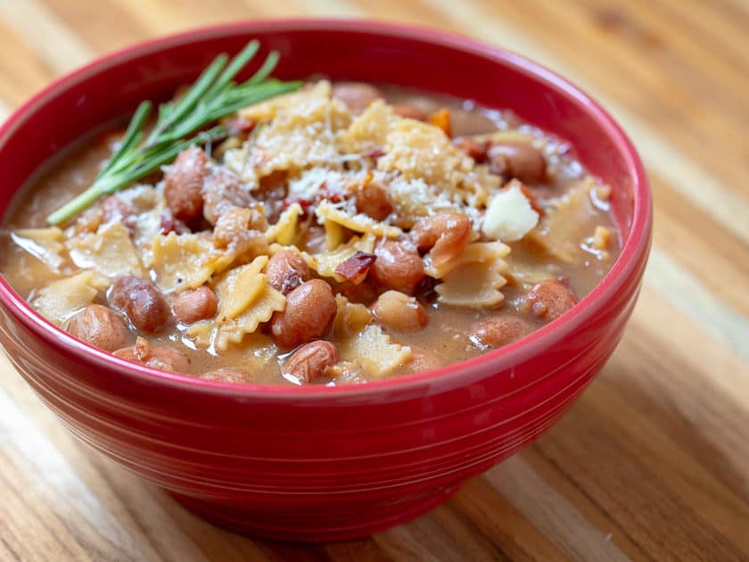A red bowl of Venetian pasta and beans garnished with a sprig of rosemary.
