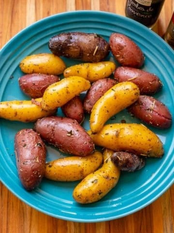 An overhead picture of a blue bowl full of multicolored fingerling potatoes, coated with herbs, with a jar of Herbes de Provence and a bottle of olive oil on the side