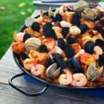 A black paella pan full of paella, bristling with shrimp, mussels, and clams, on a gray wooden table