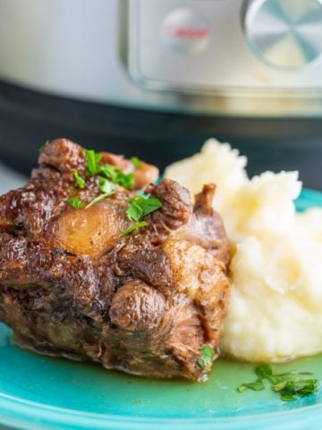 Cooked oxtail sprinkled with parsley propped up against mashed potatoes on a teal plate with the text Instant Pot Easy Braised Oxtail underneath