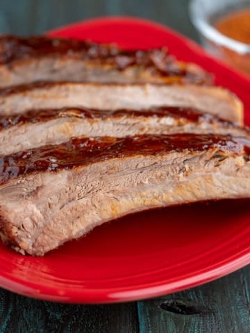 Individual spare ribs brushed with sauce on a red plate with a bowl of barbecue rub in the background