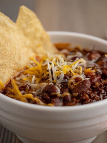 A bowl of ground beef and bean chili, with tortilla chips and cheese sprinkled on top