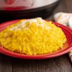 A red plate of bright yellow risotto Milanese sprinkled with grated cheese, with a napkin, spoon, and instant pot in the background