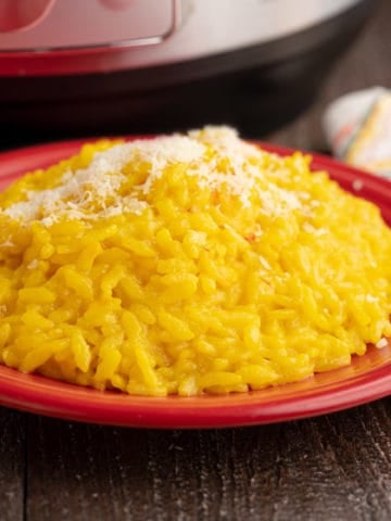 A red plate of bright yellow risotto Milanese sprinkled with grated cheese, with a napkin, spoon, and instant pot in the background