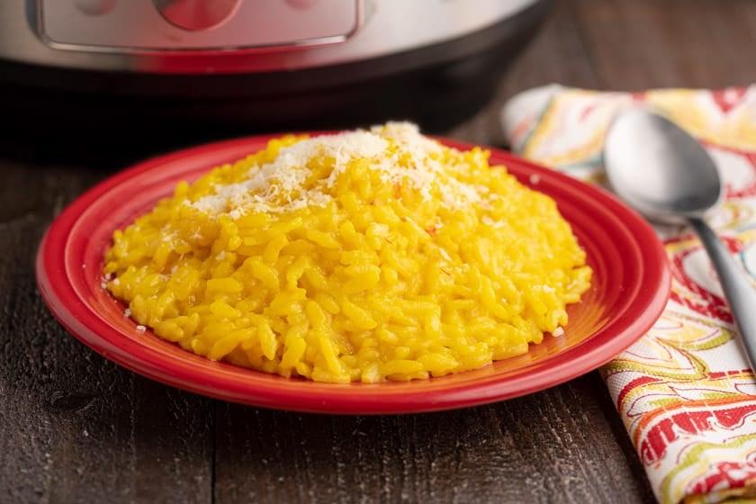 A red plate of bright yellow risotto Milanese sprinkled with grated cheese, with a napkin, spoon, and instant pot in the background