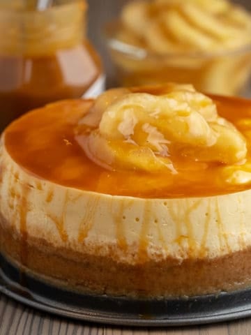 Caramel Apple Cheesecake on a wood table, in front of a jar of caramel topping and a bowl of apples with the text Instant Pot Caramel Apple Cheesecake - DadCooksDinner