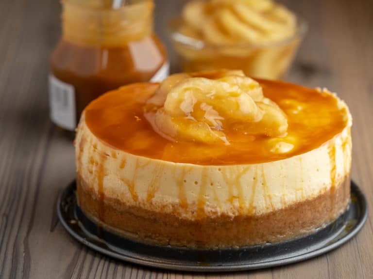 Caramel Apple Cheesecake on a wood table, in front of a jar of caramel topping and a bowl of apples with the text Instant Pot Caramel Apple Cheesecake - DadCooksDinner