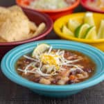 An aqua colored bowl full southwestern turkey soup, topped with shredded cabbage and cheese, with bowls of tortillas, limes, and shredded cabbage in the background, with the text Instant Pot Day-After-Thanksgiving Turkey Carcass Soup | DadCooksDinner underneath