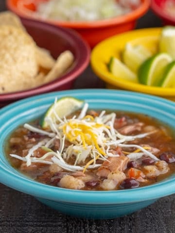 An aqua colored bowl full southwestern turkey soup, topped with shredded cabbage and cheese, with bowls of tortillas, limes, and shredded cabbage in the background, with the text Instant Pot Day-After-Thanksgiving Turkey Carcass Soup | DadCooksDinner underneath