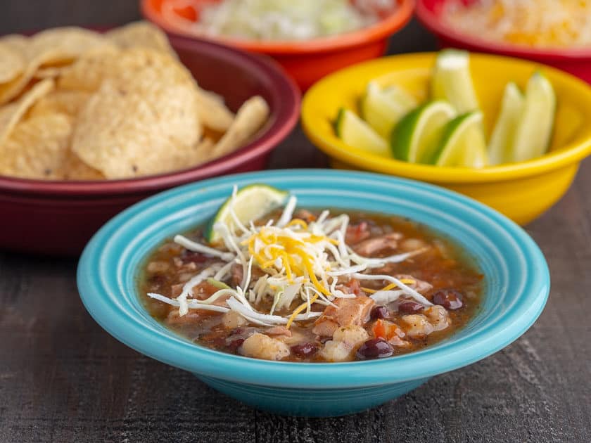 An aqua colored bowl full southwestern turkey soup, topped with shredded cabbage and cheese, with bowls of tortillas, limes, and shredded cabbage in the background, with the text Instant Pot Day-After-Thanksgiving Turkey Carcass Soup | DadCooksDinner underneath
