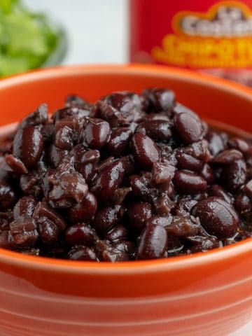 Cooked black beans in an orange bowl with a jar of chipotles behind it