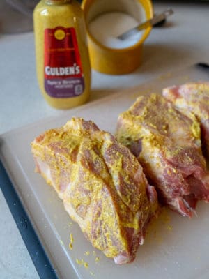 Pieces of pork shoulder roast, rubbed with mustard, on a plastic cutting board, with a bottle of mustard in the background