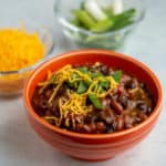 An orange bowl of turkey and bean chili, sprinkled with cheese and green onions, with a bowl of cheese and green onions in the background