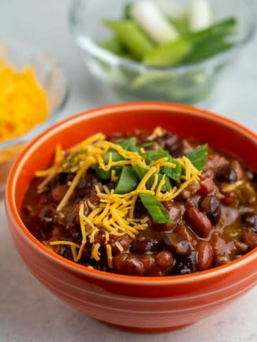 An orange bowl of turkey and bean chili, sprinkled with cheese and green onions, with a bowl of cheese and green onions in the background