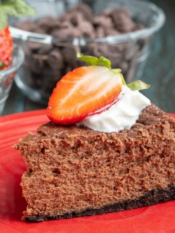 A piece of chocolate cheesecake, topped with whipped cream and a sliced strawberry, on a red plate, in front of a bowl of chocolate chips and strawberries.