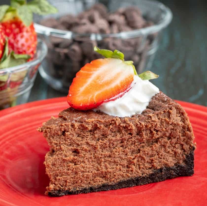 A piece of chocolate cheesecake, topped with whipped cream and a sliced strawberry, on a red plate, in front of a bowl of chocolate chips and strawberries.