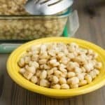 A yellow bowl of cooked hominy on a wooden tabletop, in front of a storage container of hominy