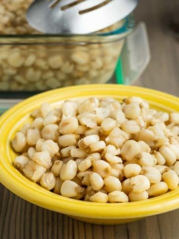 A yellow bowl of cooked hominy on a wooden tabletop, in front of a storage container of hominy