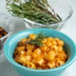 A teal bowl of pasta and chickpeas, with a sprig of rosemary on top, and a bowl of rosemary sprigs and red pepper flakes in the background.